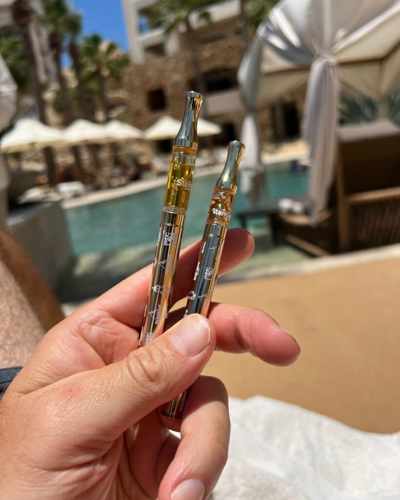 Healing Rituals for Memorial Day: A hand holding two vape pens, one filled with yellow liquid and the other clear, against a sunny poolside background.