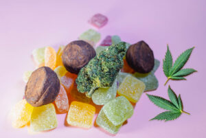 A pile of colorful gummy candies, chocolate truffles, a cannabis bud, and two cannabis leaves on a pale pink background.